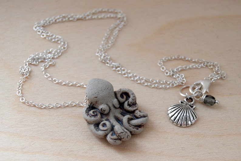 Op de afbeelding: Een zilveren ketting met een grijze keramische octopus hanger en een zilveren hanger van een schelp met een grijze edelsteen kraal.