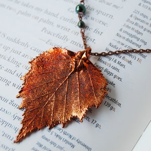 May include: A copper-colored leaf pendant necklace with a delicate chain and dark green beads. The leaf has intricate veining and a textured surface. The necklace rests on a book page with printed text.