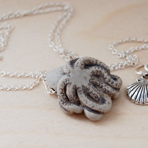 Op de afbeelding: Een zilveren ketting met een keramische octopus hanger en een bedel met een schelp en een groene edelsteen.