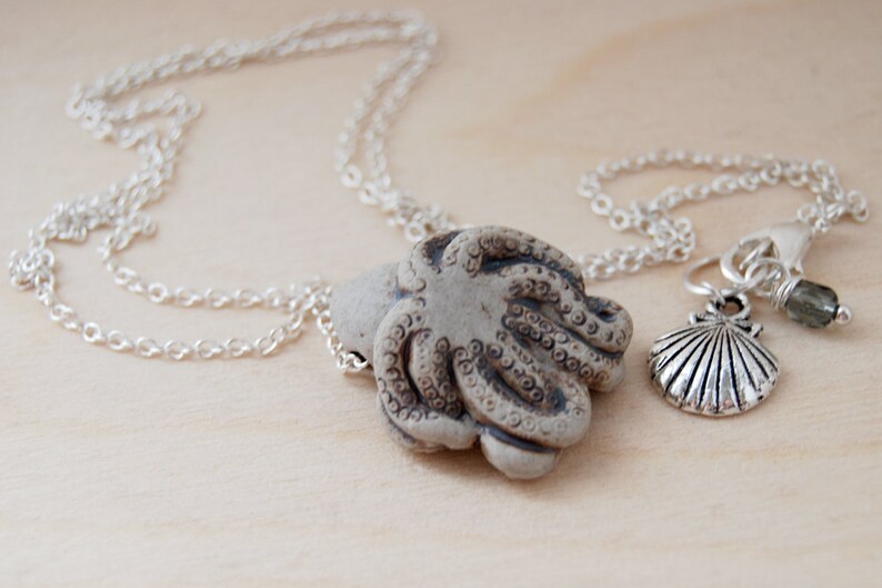 Op de afbeelding: Een zilveren ketting met een keramische octopus hanger en een bedel met een schelp en een groene edelsteen.