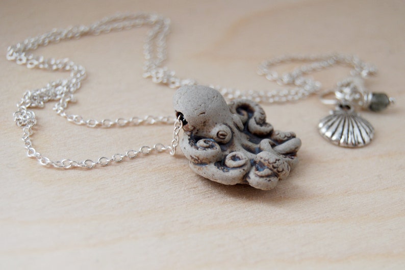 Op de afbeelding: Een zilveren ketting met een keramische octopus hanger en een zilveren schelp bedel. De octopus is grijs en heeft een getextureerd oppervlak.