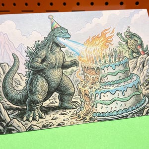 May include: A birthday card featuring a cartoon Godzilla wearing a party hat and breathing fire onto a two-tiered cake with lit candles. A turtle waves from a rocky hillside. The card has a green envelope.