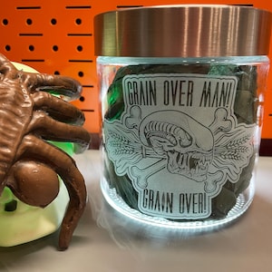 May include: A clear glass jar with a metallic lid, filled with green items, and a label that reads "GRAIN OVER MAN!". A brown insect-like sculpture is on the left. The background is orange.