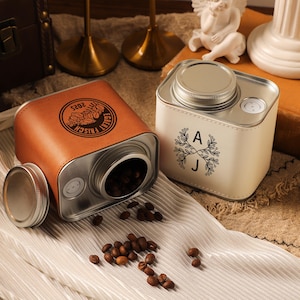 May include: Two coffee bean storage canisters. One is tan leather with a silver lid, the other is off-white with a silver lid. The tan canister has the text "Echt Frish" and "2025" printed on it. The off-white canister has the letters "A" and "J" with a floral design.
