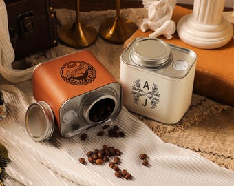 Pode incluir: Dois recipientes decorativos para grãos de café. Um é de couro castanho com tampa prateada, o outro é branco sujo com tampa prateada. O recipiente castanho tem o texto "Echt Frish" e "2025". O recipiente branco sujo tem as letras "A" e "J" com um desenho floral.