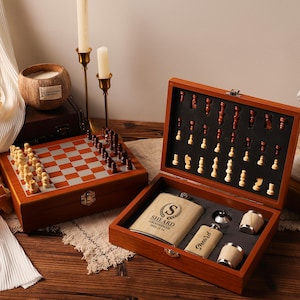 May include: A wooden chess set with a chessboard and chess pieces, alongside a wooden box containing a flask, shot glasses, and a flask. The chess set has a brown and silver checkered board. The box has the name "Sheard" on it.