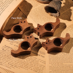 May include: Four wooden cat napkin rings. The rings are carved in the shape of a cat with a hole in the middle for a napkin. The rings are a dark brown colour and have a smooth finish.