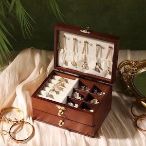 May include: A dark brown wooden jewelry box with a hinged lid and a pull-out drawer. The interior features a cream-colored velvet lining with hooks for necklaces, padded slots for rings, and several small compartments for storing various accessories.