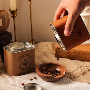 May include: A brown leather-covered flask is pouring coffee beans into a small wooden bowl. A square coffee storage tin, labelled "Barnaby Wendt Coffee Storage", is also present. The scene is set on a beige cloth.