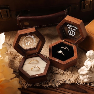 Könnte beinhalten: Zwei offene sechseckige Holzringboxen mit Glasdeckeln, die Ringe auf einem cremefarbenen und schwarzen Samtinnenraum zeigen. Die Boxen sind aus dunklem Holz und stehen auf einer strukturierten Oberfläche. Eine kleine weiße Statue befindet sich im Vordergrund.