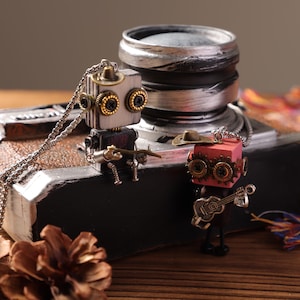 Puede incluir: Dos collares con colgantes de robots. Un robot es plateado y negro, el otro es rojo y negro. El robot plateado tiene una cadena y está sentado en un objeto negro y marrón. El robot rojo toca la guitarra y lleva un sombrero.