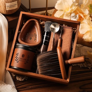 Puede incluir: Un juego de caja de madera que contiene varios artículos. El juego incluye un cuenco de madera pequeño, un tarro con el texto "CAESAR 2025.07.24", un cepillo, una cuchara y una toalla pequeña. Los artículos son marrones y beige, con una estética natural.