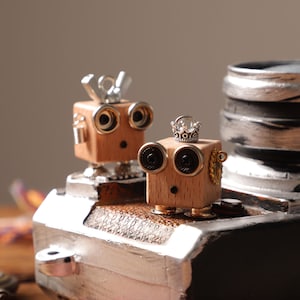 May include: Two small wooden robot figures with round eyes and metal details. One wears a silver crown, the other has a wing nut on top. They are positioned on a metallic surface against a dark backdrop.