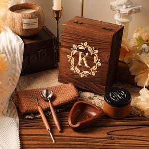 May include: A wooden box with a decorative "K" and floral design, a coconut shell candle, and a small jar with a black lid. Also included are a small wooden bowl, a brown washcloth, and two wooden-handled metal tools. The items are arranged on a wooden surface.