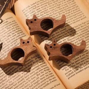 May include: Three wooden cat-shaped ring holders. Each ring holder is carved with a cat face and has a ring slot in the middle. The ring holders are on a book with text about the Wright brothers.