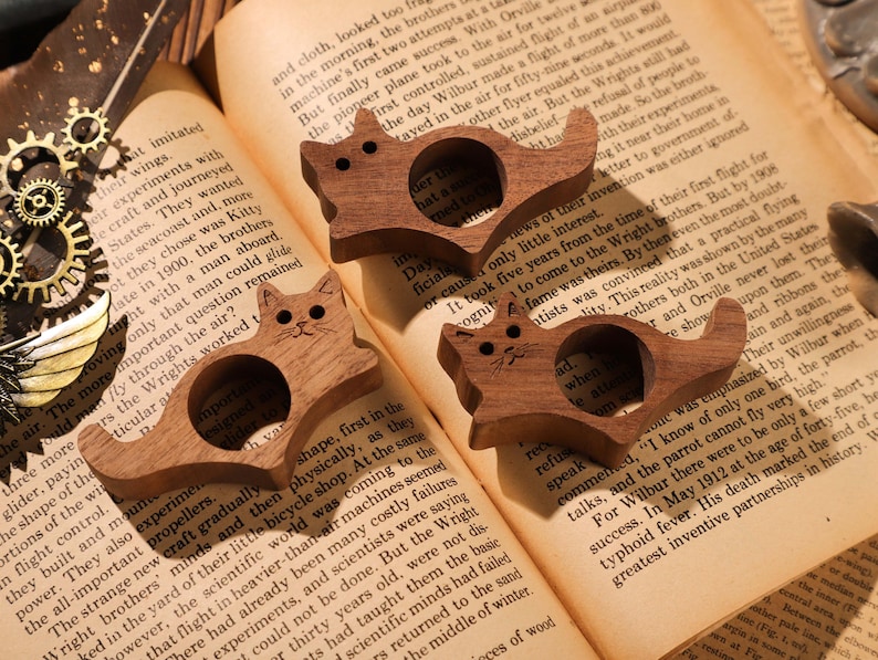 May include: Three wooden cat-shaped ring holders. Each ring holder is carved with a cat face and has a ring slot in the middle. The ring holders are on a book with text about the Wright brothers.