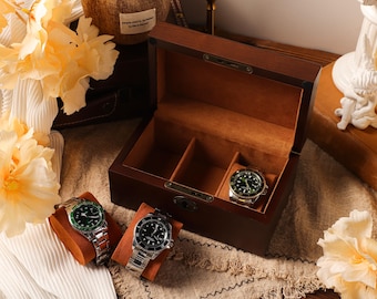 Op de afbeelding: Een donkerbruine houten horlogedoos met drie compartimenten, waarvan één met een horloge. Twee extra horloges met groene en zwarte wijzerplaten rusten op oranje horlogekussens. De horloges hebben zilveren banden. De doos is open en onthult een bruin interieur.
