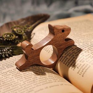 Puede incluir: Un soporte de anillo de madera con forma de ardilla, sentado sobre un libro con una cubierta marrón. El soporte de anillo está hecho de madera marrón oscura y tiene un acabado natural.