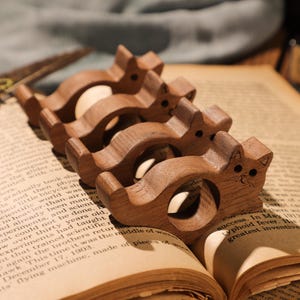 May include: Four wooden cat-shaped napkin rings, each with a different size and a carved face. The rings are arranged in a row on a book.