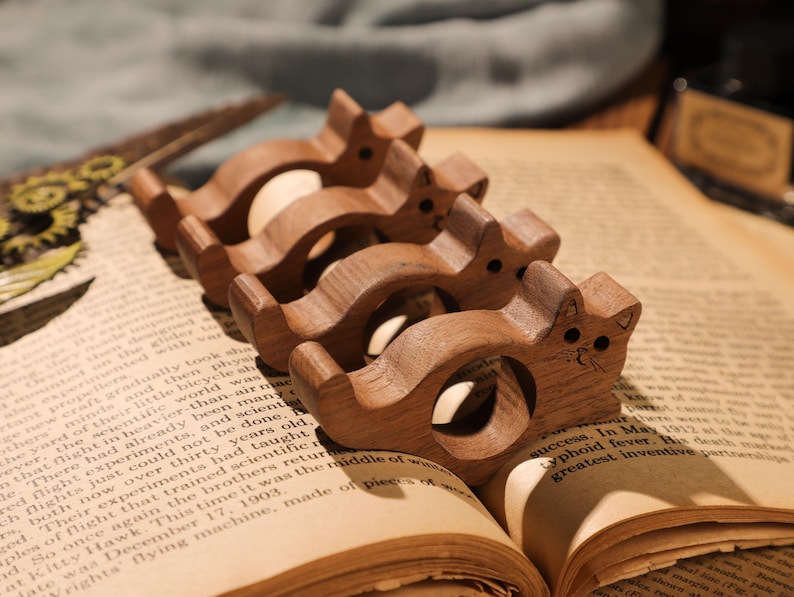 May include: Four wooden cat-shaped napkin rings, each with a different size and a carved face. The rings are arranged in a row on a book.