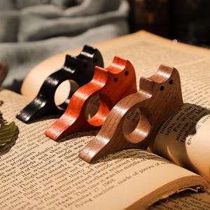 May include: Three wooden bookends shaped like birds. The bookends are in different shades of brown and red. The bookends are sitting on top of an open book.
