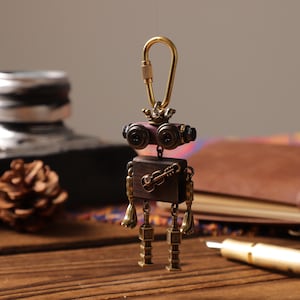 Puede incluir: Un llavero de robot de estilo steampunk con un mosquetón dorado. El robot presenta un cuerpo de madera oscura, patas de latón y una cabeza con dos lentes oscuras. Una pequeña guitarra está grabada en el cuerpo. El llavero cuelga sobre un fondo borroso.