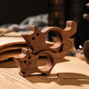 May include: Two wooden cat-shaped ring holders, stacked on top of each other. The holders are made of dark brown wood and have a smooth, polished finish.