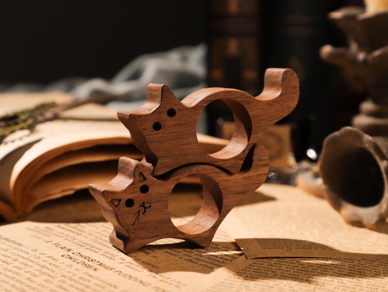 May include: Two wooden cat-shaped ring holders, stacked on top of each other. The holders are made of dark brown wood and have a smooth, polished finish.