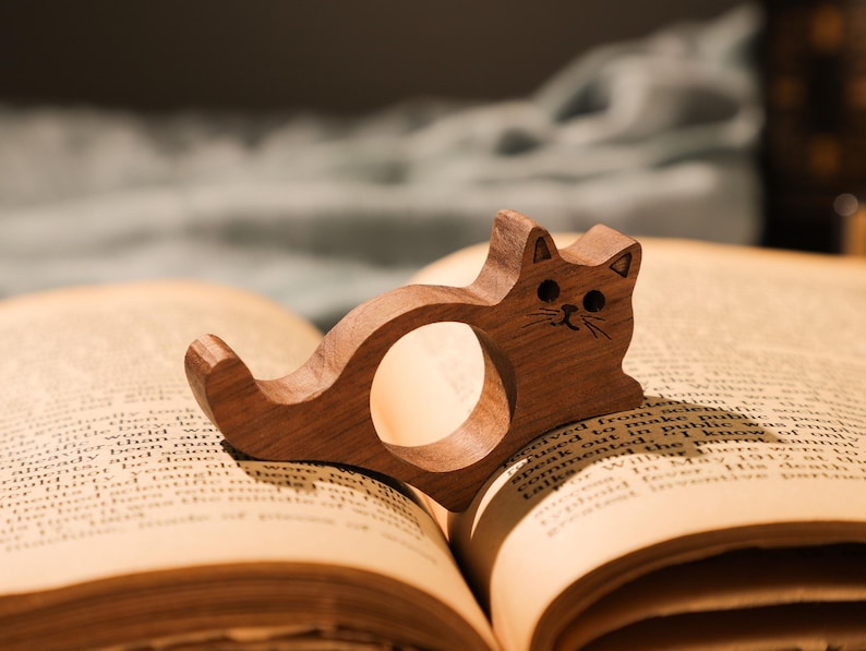 May include: A wooden cat bookmark with a carved face and a ring to hold a page. The bookmark is resting on an open book.