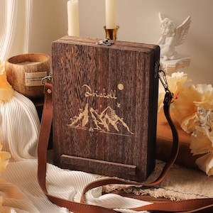 May include: A wooden box bag with a brown leather strap. The bag features a mountain and sun design with the word "Driscoll" engraved on the front. The bag has a metal clasp closure.