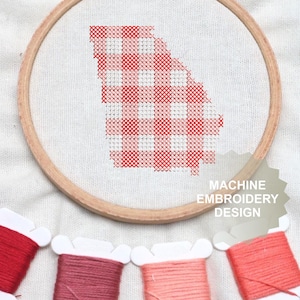 May include: A wooden embroidery hoop containing white fabric with a red and white gingham pattern stitched in the shape of the state of Georgia. Below the hoop are four plastic bobbins wrapped in red and pink embroidery thread. Text reads MACHINE EMBROIDERY DESIGN.