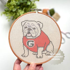 Può includere: Ricamo a punto croce di un bulldog con una maglia rossa con una "G" nera. Il ricamo è posto in un telaio di legno. Il bulldog è seduto con la lingua di fuori. Lo sfondo è sfocato.