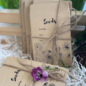 May include: Several seed packets made of brown paper are tied with twine. The word "Seeds" is written in a script font on the front of each packet, along with space for variety, from, and date. A purple flower rests on one packet.