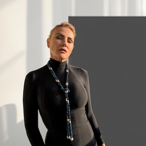 May include: A woman wearing a long, dark blue beaded necklace with gold and white accents. The necklace is draped over a black turtleneck dress. The background is a mix of white and dark gray.