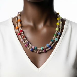 May include: A multi-strand necklace featuring colorful beaded spheres. The necklace has a neutral-colored cord and is composed of multiple strands. The beads are in a variety of colors, including red, orange, yellow, green, blue, and purple.