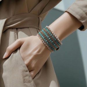 Puede incluir: Una pulsera ancha con cuentas turquesas, doradas y blancas. La pulsera se lleva en la muñeca, mostrando el intrincado patrón y la combinación de colores. El diseño incluye múltiples filas de cuentas, creando un accesorio texturizado y llamativo. Circunferencia aproximada de 17 cm.