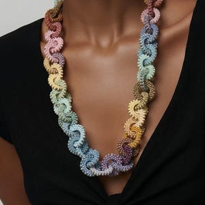 Pode incluir: Um colar multicolorido com um design em espiral. O colar apresenta um gradiente de cores pastel, incluindo rosa, amarelo, azul e roxo. É feito de pequenas contas texturizadas e usado sobre uma blusa preta.