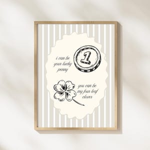 May include: A framed print with a light beige background and vertical stripes. The print features a coin and a four-leaf clover illustration, with the text "I can be your lucky penny" and "you can be my four leaf clover". The frame is a light wood color.