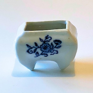May include: A small white ceramic planter with a blue floral design. The planter has four small feet and is ideal for a small succulent or other plant.