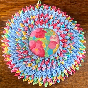 Op de afbeelding: Een handgemaakte, cirkelvormige onderzetter met een bloemenontwerp. De onderzetter heeft een centraal, rond stoffen stuk met roze bloemen en groene bladeren. De buitenste ring is gemaakt van blauwe, roze, gele en groene stoffen stroken, waardoor een gelaagd, getextureerd effect ontstaat. Er is een lus bevestigd om op te hangen.