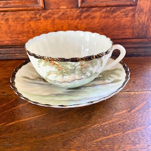 Peut inclure: Une tasse à thé et une soucoupe blanches avec un bord noir et doré. La tasse et la soucoupe sont décorées d'une scène de paysage verte et dorée.