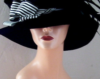 May include: A black hat featuring a wide brim and a structured crown. The hat is adorned with a large, layered bow made of black and white striped fabric on the left side, complemented by dark, slender feathers extending toward the right side.