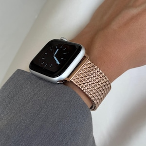 Op de afbeelding: Een zilveren Apple Watch met een roségouden Milanese-band. De wijzerplaat van het horloge is zwart met witte wijzers en cijfers.