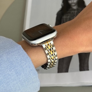 Puede incluir: Un Apple Watch plateado y dorado con una banda de metal. La esfera del reloj es negra y la banda está hecha de eslabones de metal entrelazados.
