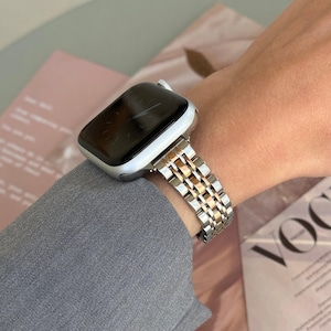 May include: A silver and gold Apple Watch with a metal band. The watch face is black and the band is made of linked metal pieces.