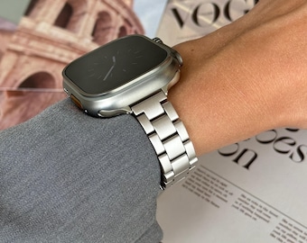 May include: A silver Apple Watch with a square face and a silver metal band. The watch is on a person's wrist.