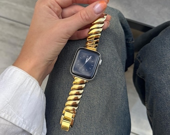 May include: A gold-colored watch band with a unique, textured design is attached to a silver smartwatch. The watch face displays a dark blue screen with white hands. The band is held by a person wearing a white shirt and blue jeans.