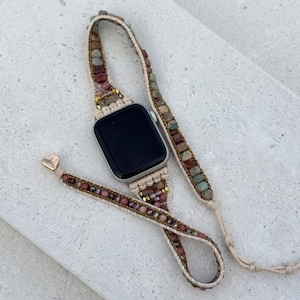 Pode incluir: Uma pulseira de smartwatch com design envolvente e contas de pedra multicoloridas em tons de rosa, castanho e cinzento, tecidas num cordão claro. A pulseira inclui um pequeno pendente metálico em forma de coração na extremidade.