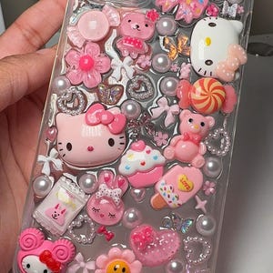 Puede incluir: Una funda transparente para teléfono decorada con adornos rosas, blancos y plateados. La funda presenta personajes de Hello Kitty, lazos, corazones, flores y caramelos.