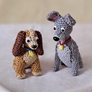 Könnte beinhalten: Zwei gehäkelte Hundefiguren. Einer ist braun und braun mit Schlappohren, einem blauen Halsband und einem gelben Anhänger. Der andere ist grau mit einem roten Halsband und einem gelben Anhänger. Beide haben schwarze Nasen und große, ausdrucksstarke Augen.